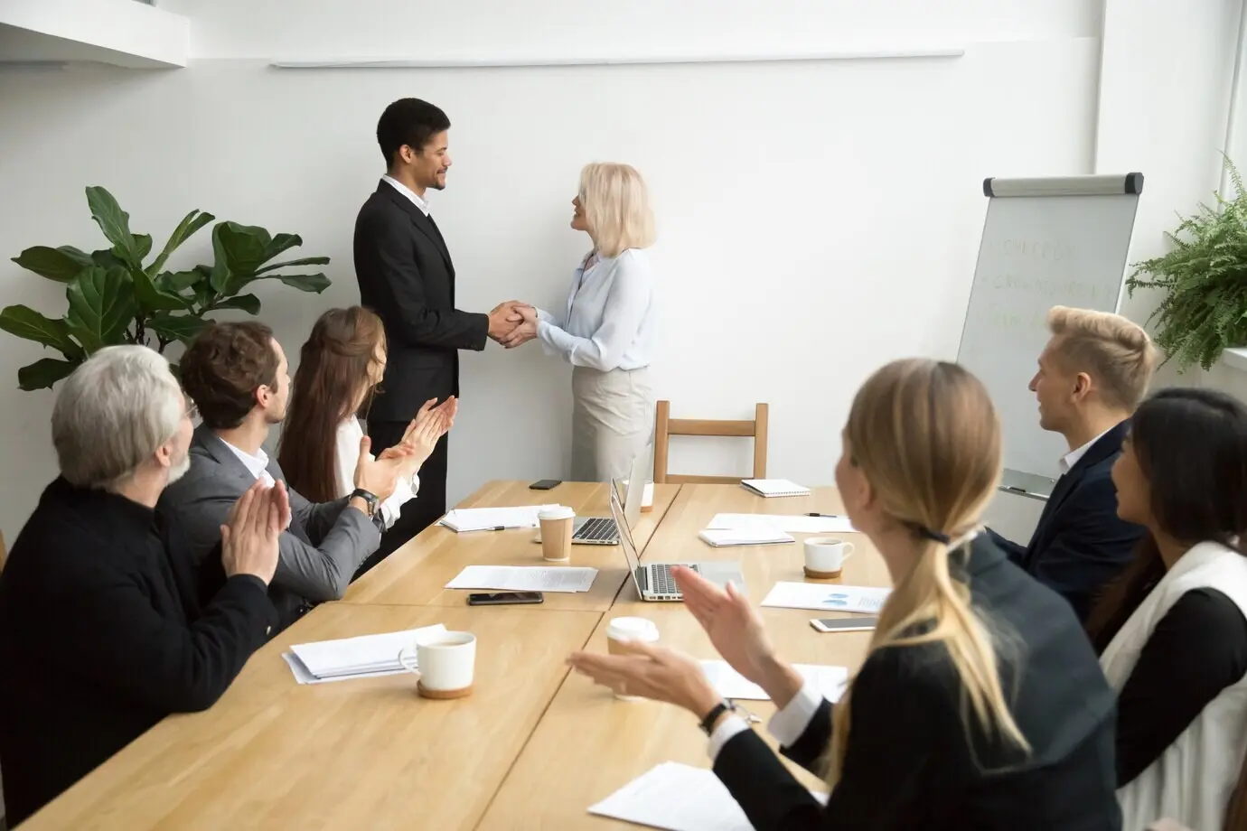 Een senior vrouwelijke manager promoveert een Afrikaanse medewerker en schudt de medewerker de hand, terwijl het team applaudisseert.