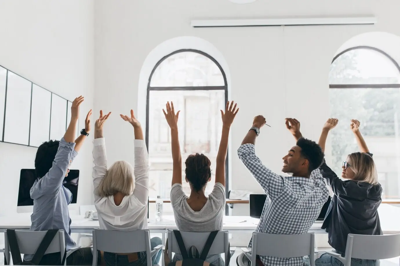 Een vrouw met blond haar in een formele blouse zwaait met haar handen en zit tussen collega’s in een grote, lichte conferentiezaal. Foto van achteren van vermoeide managers die zich uitrekken tijdens een vergadering op kantoor.